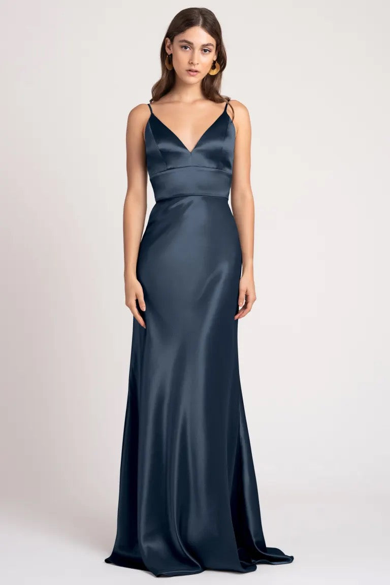 A woman in an elegant, dark blue Brenna bridesmaid dress made of satin back crepe fabric by Jenny Yoo stands against a neutral background from Bergamot Bridal.
