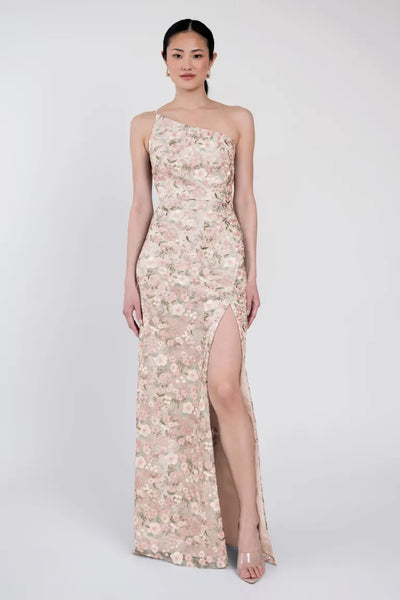 A woman stands wearing the Bristol Jenny Yoo Bridesmaid Dress from Bergamot Bridal, an Enchanted Floral, one-shoulder gown with a high slit, set against a plain white background.