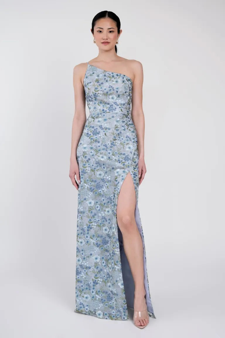 A person wearing the enchanted floral, one-shoulder, floor-length Bristol gown with a high slit stands against a plain background. The Jenny Yoo Bridesmaid Dress by Bergamot Bridal gracefully drapes around them, catching the light with its delicate details.