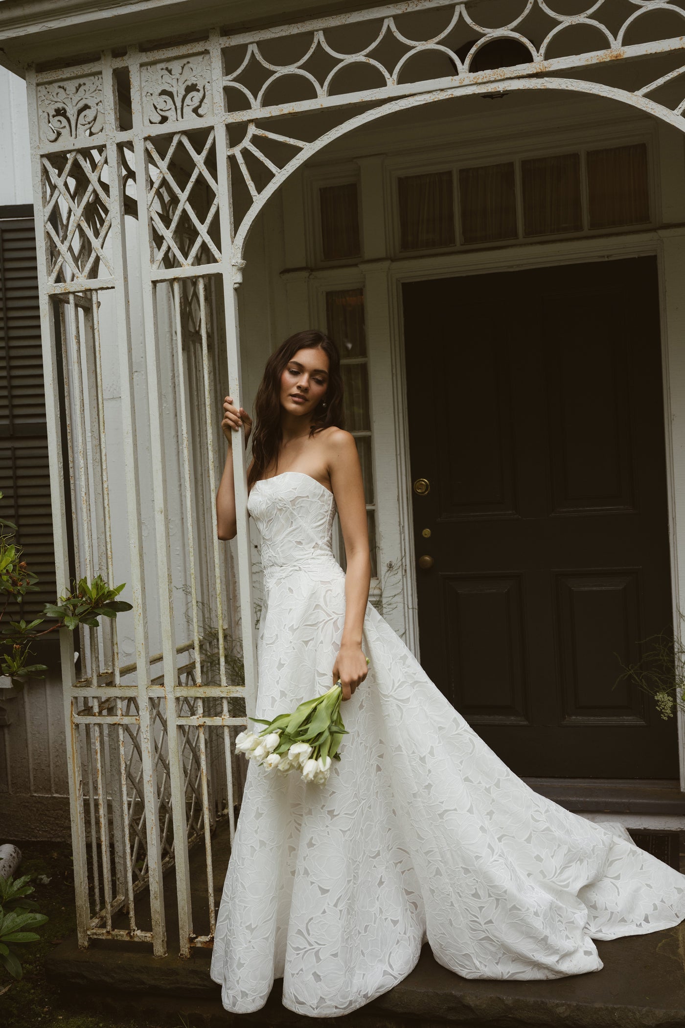 A woman in the strapless white "Brody by Jenny Yoo" gown from Bergamot Bridal with a subtle floral motif stands by an ornate gate, holding white tulips, in front of a dark wooden door.