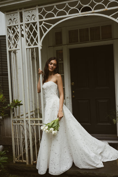 A woman in the strapless white "Brody by Jenny Yoo" gown from Bergamot Bridal with a subtle floral motif stands by an ornate gate, holding white tulips, in front of a dark wooden door.