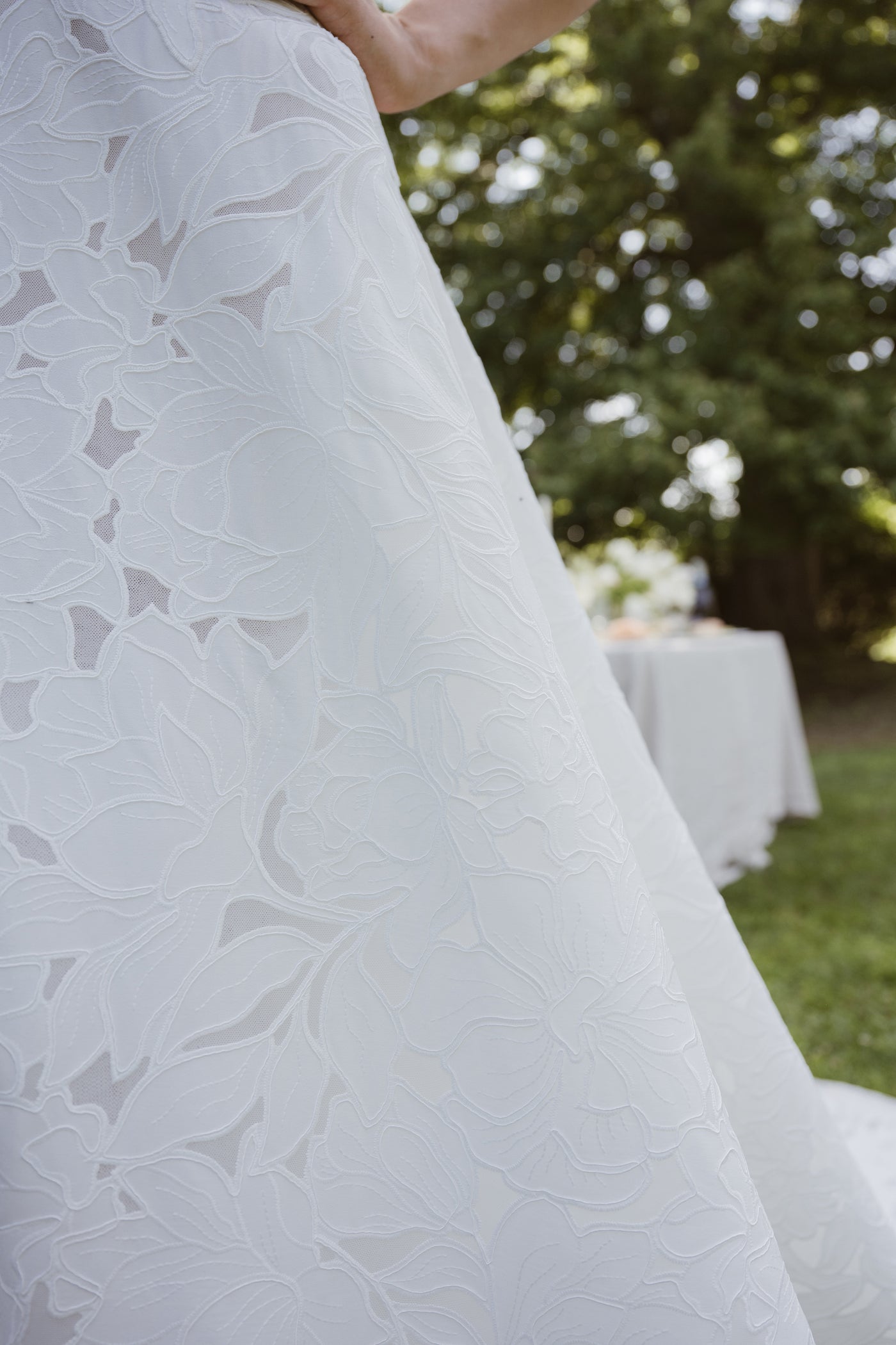 Close-up of the Brody by Jenny Yoo gown from Bergamot Bridal, featuring a delicate floral motif, photographed outdoors among grass, trees, and a table with a white cloth.