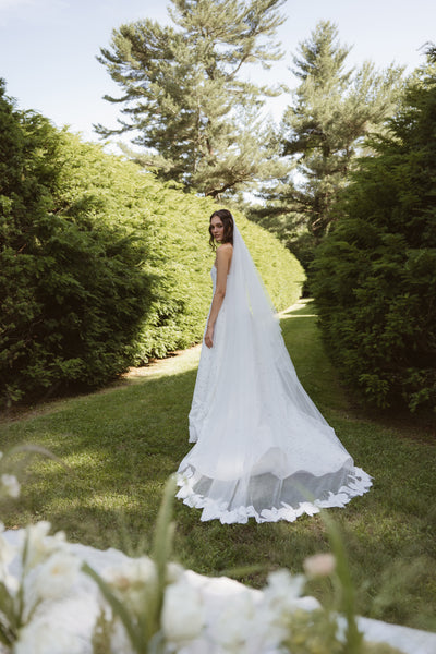 A bride wears the Brody by Jenny Yoo gown from Bergamot Bridal, featuring a delicate floral motif and long veil, standing on a grassy path between tall green hedges and glancing over her shoulder.