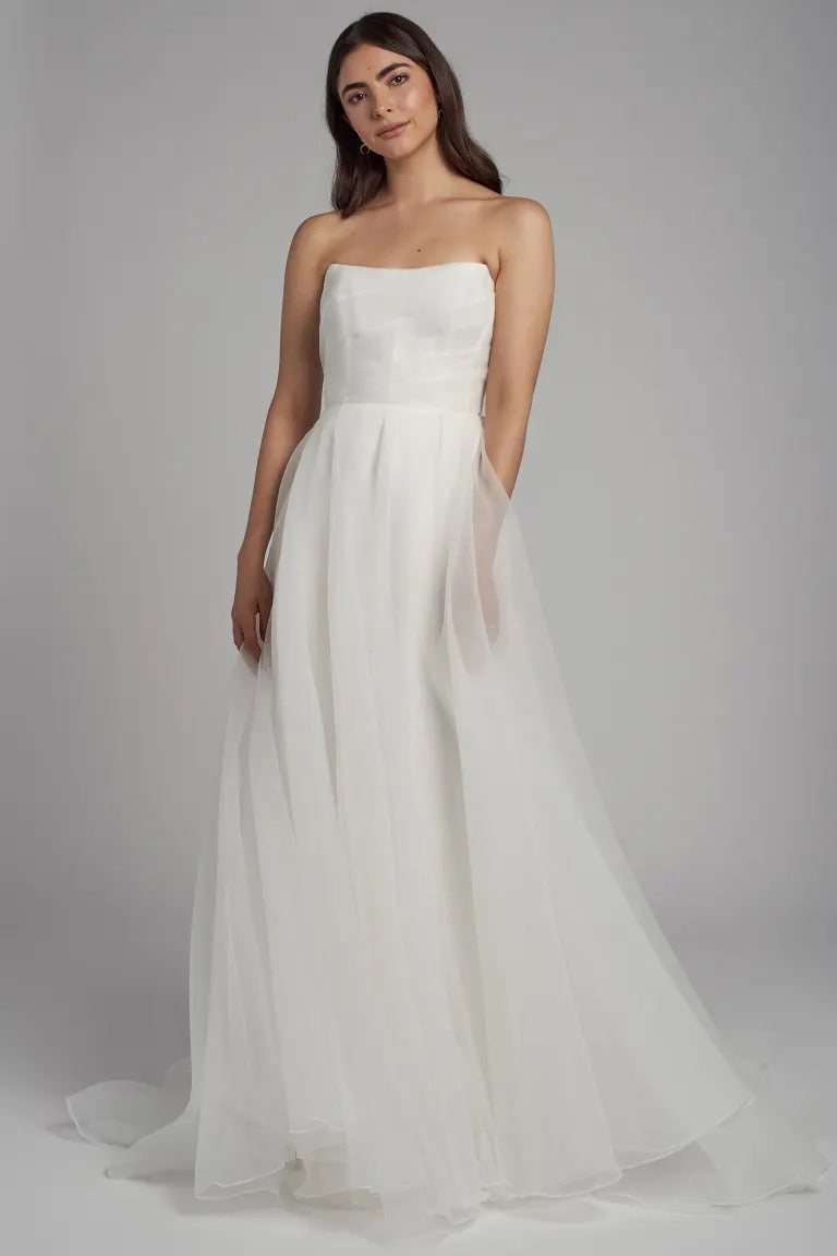 A woman poses in the Bronte Wedding Dress by Jenny Yoo for Bergamot Bridal; the strapless white gown features an A-line skirt and pebbled organza, adding elegance to her enchanting look against a gray backdrop.