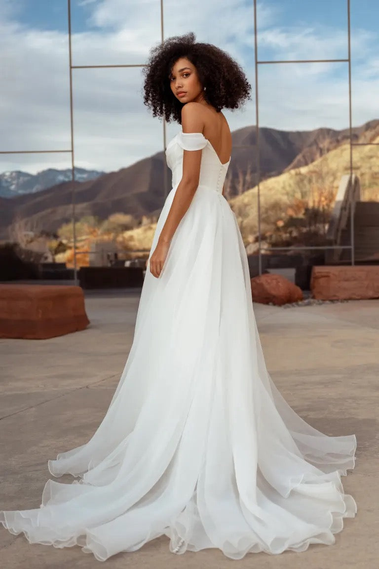 A woman in the Bronte Wedding Dress by Jenny Yoo from Bergamot Bridal stands outdoors, framed by mountains and a reflective building. The white off-the-shoulder A-line gown's pebbled organza softly catches the light, adding texture to the elegant scene.