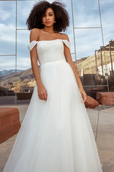 A woman in the Bronte Wedding Dress by Jenny Yoo, available at Bergamot Bridal, stands outdoors in an off-the-shoulder white bridal gown with a pebbled organza texture, the clear sky reflecting on glass walls behind her.