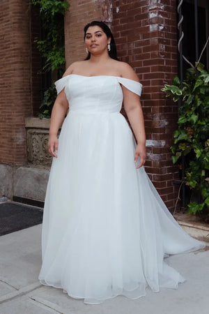 A woman in the Bronte Wedding Dress by Jenny Yoo, sold by Bergamot Bridal, poses gracefully in front of a brick wall embellished with lush greenery.