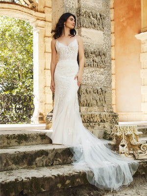 Wearing the elegant Beaded Fit & Flare Gown with Soft Tulle Skirt by Bergamot Bridal, a woman stands on stone steps beside an ornate column, surrounded by greenery and an archway in the background.