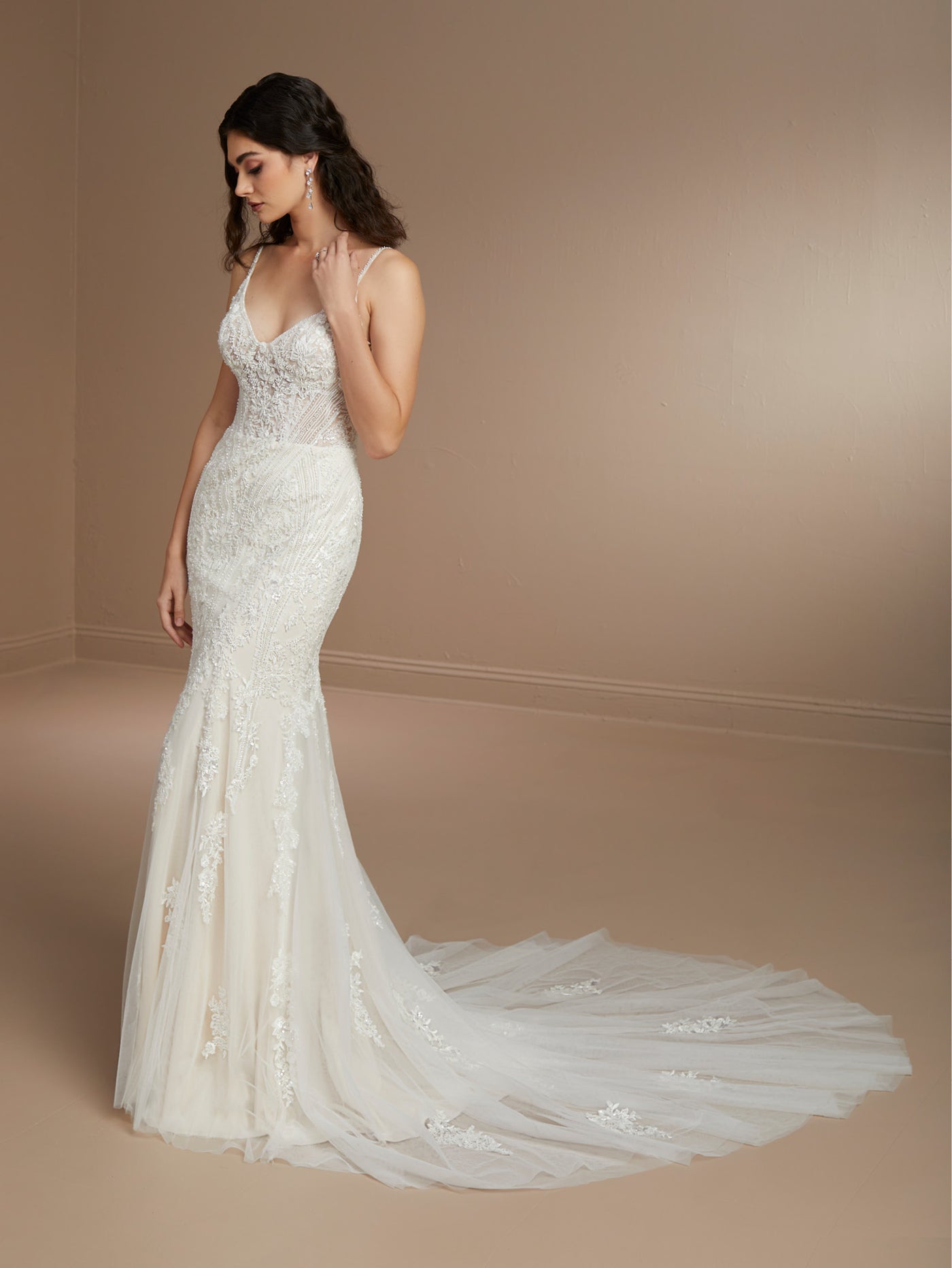 A woman gracefully stands in a neutral-colored room wearing the Beaded Fit & Flare Gown with Soft Tulle Skirt by Christina Wu from Bergamot Bridal, the dress's elegant train pooling on the floor. The stunning gown features a sweetheart neckline and intricate beaded lace detailing.