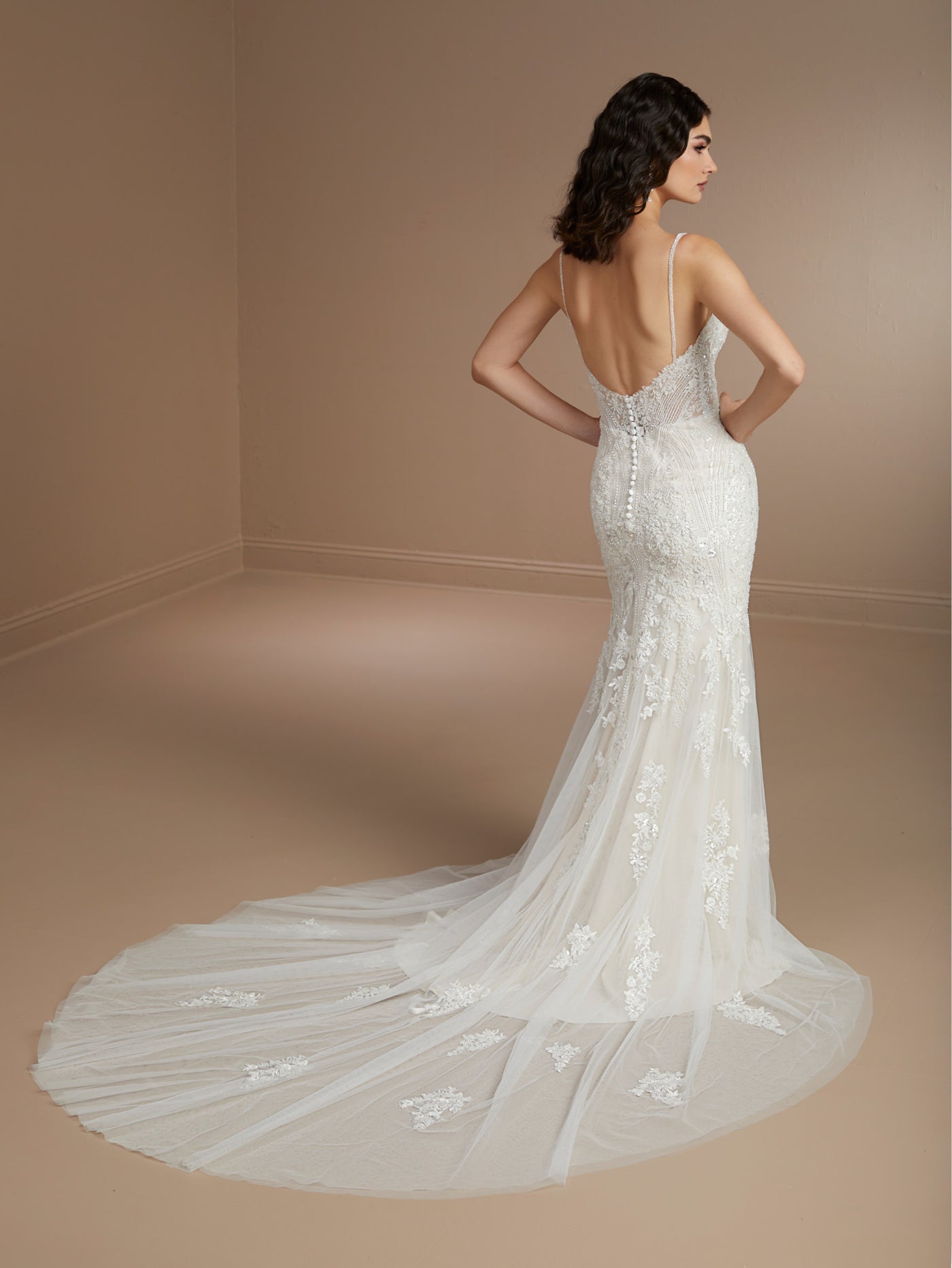 A woman dressed in the Beaded Fit & Flare Gown with Soft Tulle Skirt by Christina Wu from Bergamot Bridal stands in a minimalist room, facing away.