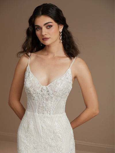 A woman with long dark hair wears the Beaded Fit & Flare Gown with Soft Tulle Skirt by Christina Wu from Bergamot Bridal, featuring a detailed white lace design with thin straps and intricate beading. The gown elegantly highlights her silhouette as she stands against a neutral background.