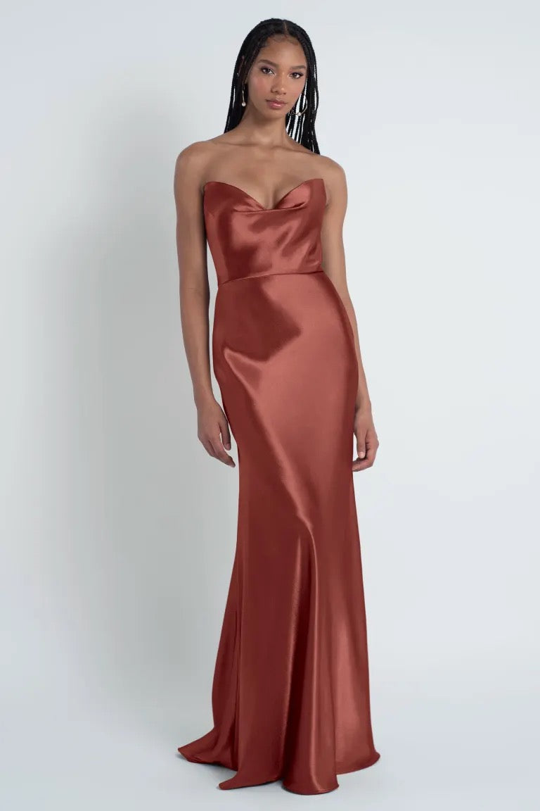 A woman stands against a plain background wearing a strapless, floor-length satin Camilla - Bridesmaid Dress by Jenny Yoo in a rich, rust color. The dress features a bias cut skirt and a sculpted cowl sweetheart neck, adding an elegant touch to her sophisticated look. This elegant piece is from the collection of Bergamot Bridal.