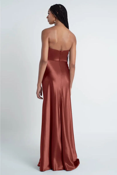 A woman with braided hair is wearing a strapless, floor-length, rust-colored satin gown. The Camilla - Bridesmaid Dress by Jenny Yoo from Bergamot Bridal features a sculpted cowl sweetheart neck and bias cut skirt. She stands with her back facing the camera against a plain white background.