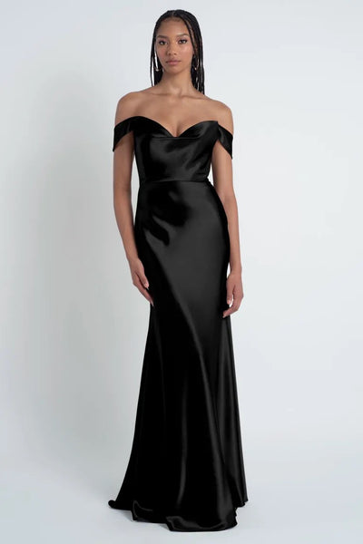 A person stands wearing a floor-length black Camilla - Bridesmaid Dress by Jenny Yoo, posing against a plain white background.