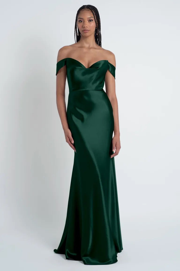 A woman stands wearing a dark green, off-the-shoulder Camilla - Bridesmaid Dress by Jenny Yoo from Bergamot Bridal with a sculpted cowl sweetheart neck and bias cut skirt, against a plain white background.