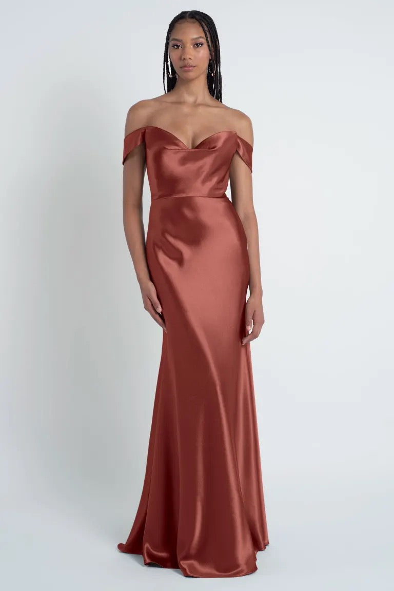 A woman in an off-the-shoulder, floor-length satin Camilla - Bridesmaid Dress by Jenny Yoo in a rust color, featuring a sculpted cowl sweetheart neck and bias cut skirt, stands against a plain white background.