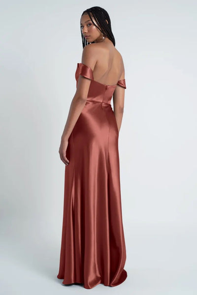 A woman wearing a satin, off-the-shoulder, burnt orange Camilla - Bridesmaid Dress by Jenny Yoo with a bias cut skirt stands facing sideways, looking over her shoulder. The background is plain white.