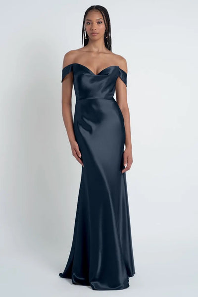 A woman stands wearing an off-the-shoulder navy blue satin Camilla - Bridesmaid Dress by Jenny Yoo from Bergamot Bridal with a sculpted cowl sweetheart neck. She has long hair styled in braids.