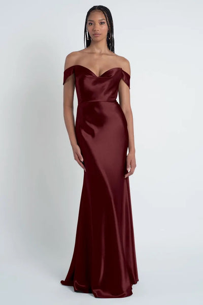 A woman stands against a plain background, wearing a floor-length, off-the-shoulder, dark red satin Camilla - Bridesmaid Dress by Jenny Yoo from Bergamot Bridal. The sculpted cowl sweetheart neck adds elegance to the ensemble. She has long braided hair and a neutral expression.