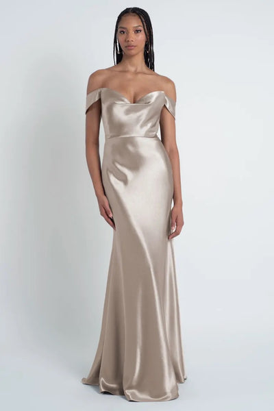 A woman in an off-the-shoulder champagne-colored satin gown with a sculpted cowl sweetheart neck and bias cut skirt, the Camilla - Bridesmaid Dress by Jenny Yoo from Bergamot Bridal, stands against a plain white background.