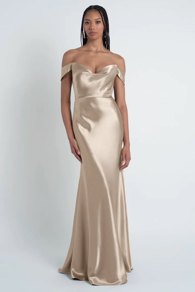 Person wearing a long, off-the-shoulder Camilla - Bridesmaid Dress by Jenny Yoo in champagne satin with a bias cut skirt, standing against a plain white background.