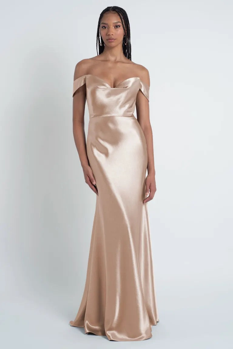 Person in a silky, off-the-shoulder, champagne-colored Camilla - Bridesmaid Dress by Jenny Yoo from Bergamot Bridal with a bias cut skirt and sculpted cowl sweetheart neck standing against a plain white background.