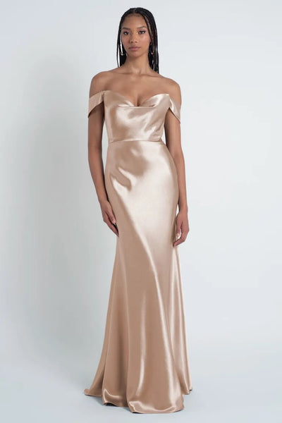 Person in a silky, off-the-shoulder, champagne-colored Camilla - Bridesmaid Dress by Jenny Yoo from Bergamot Bridal with a bias cut skirt and sculpted cowl sweetheart neck standing against a plain white background.