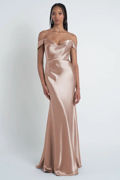 A person stands wearing a floor-length, off-shoulder Camilla - Bridesmaid Dress by Jenny Yoo in a metallic champagne color with a bias cut skirt against a light gray background, from Bergamot Bridal.