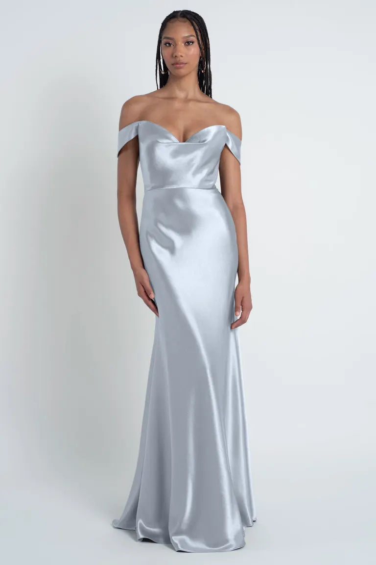 A woman is wearing a Camilla - Bridesmaid Dress by Jenny Yoo from Bergamot Bridal, featuring detachable off-the-shoulder sleeves. Its floor-length satin fabric shimmers elegantly as she stands against a plain white background.