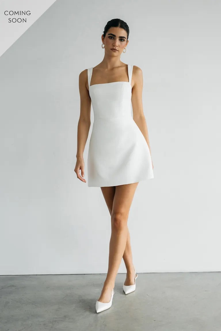 A woman wearing the chic Camille white dress by Bergamot Bridal, paired with elegant white heels, stands against a plain gray backdrop. The phrase "Coming Soon" teases the arrival of our stunning Jenny Yoo design crafted from Luxe Faille.