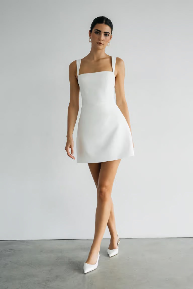 A woman stands indoors against a plain white wall, wearing the Bergamot Bridal Camille - Jenny Yoo Little White Dress with pointed-toe heels—ideal for a bridal shower or elegant event.