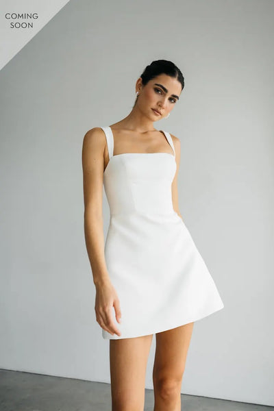 A woman in a sleeveless Camille dress by Bergamot Bridal stands against a plain backdrop. The Luxe Faille fabric adds elegance to the Jenny Yoo Little White Dress, with "Coming Soon" text in the corner.