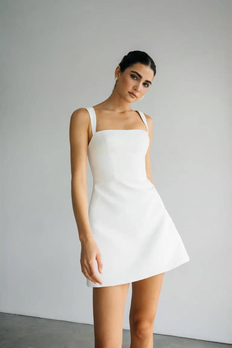 A person stands against a plain background wearing the Camille - Jenny Yoo Little White Dress by Bergamot Bridal, featuring a sleeveless, square-neck design—perfect for a bridal shower—and looks slightly to the side.