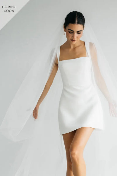 A person wearing the Camille - Jenny Yoo Little White Dress by Bergamot Bridal, paired with a sheer veil, perfect for a chic wedding after-party or bridal shower event.