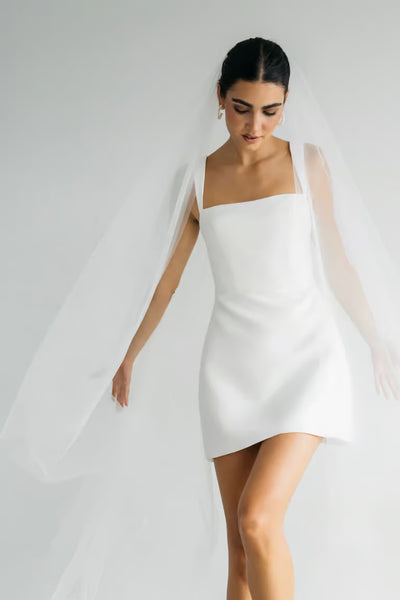 A woman in the Camille – Jenny Yoo Little White Dress from Bergamot Bridal, paired with a long white veil, stands against a plain light background—perfect for your next bridal shower or little white dress occasion.