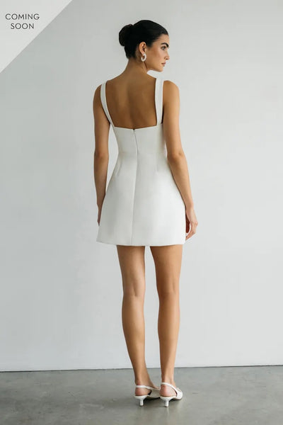 Wearing the Camille - Jenny Yoo Little White Dress by Bergamot Bridal, a woman in heels stands with her back to the camera against a plain wall, ready for the wedding after-party.