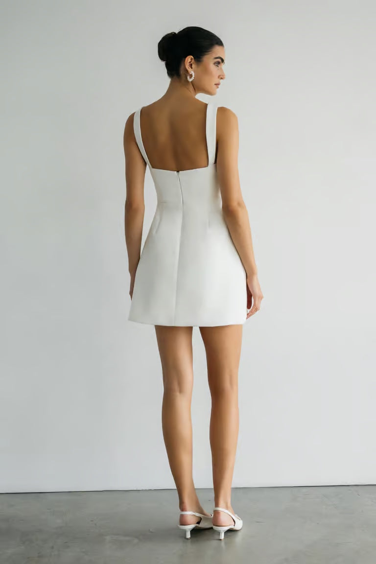 A woman stands against a plain wall wearing the Camille - Jenny Yoo Little White Dress by Bergamot Bridal, paired with white heels and her hair in a neat bun—a chic look, perfect for a bridal shower.