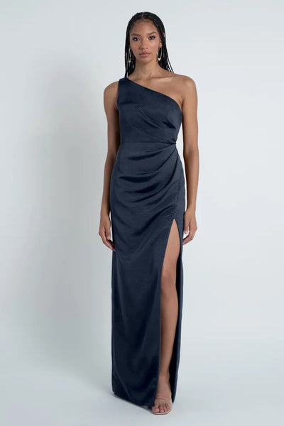 A woman poses in a one-shoulder Jenny Yoo Bridesmaid Dress made of Luxe Faille fabric, featuring a high leg slit and a navy blue hue from Bergamot Bridal.