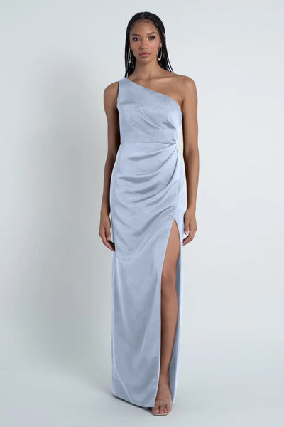 Woman in a Carolina - Jenny Yoo Bridesmaid Dress, featuring a Luxe Faille fabric and one-shoulder neckline, with a thigh-high slit standing barefoot against a plain background.