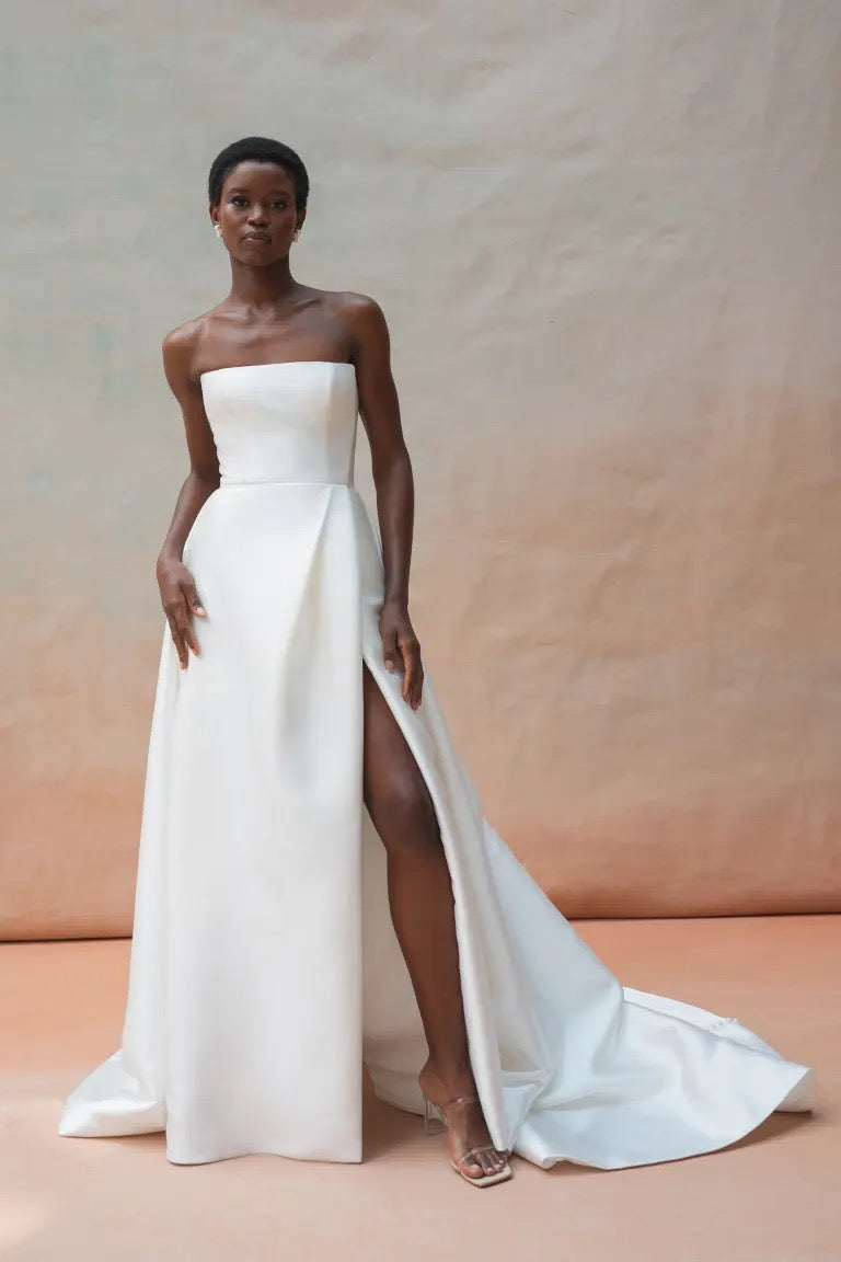 A person stands wearing the Catherine Wedding Dress by Jenny Yoo, crafted from sleek Mikado fabric with a high slit, embodying modern Hollywood glamour as they pose against a neutral backdrop. Available at Bergamot Bridal.