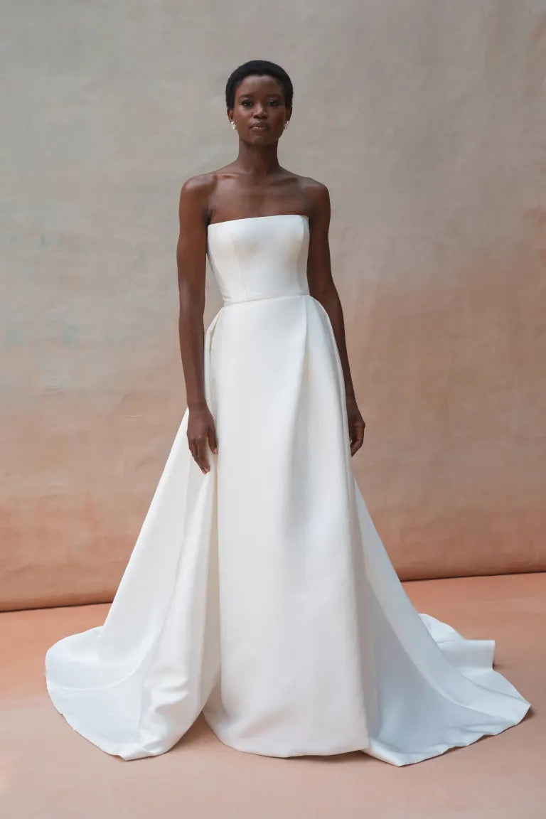 A person stands wearing the Catherine Wedding Dress by Jenny Yoo, a strapless white gown made of sleek Mikado fabric featuring a dramatic flared skirt, against a plain beige background. This stunning piece is from Bergamot Bridal.