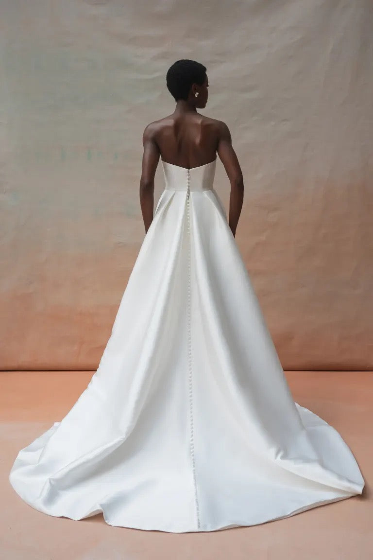 A person stands facing away, wearing the Catherine Wedding Dress by Jenny Yoo from Bergamot Bridal. The strapless gown is crafted from luxurious Mikado fabric and features a long train with elegant button detailing down the back, embodying Hollywood glamour. The background is a gradient of soft beige and light peach.