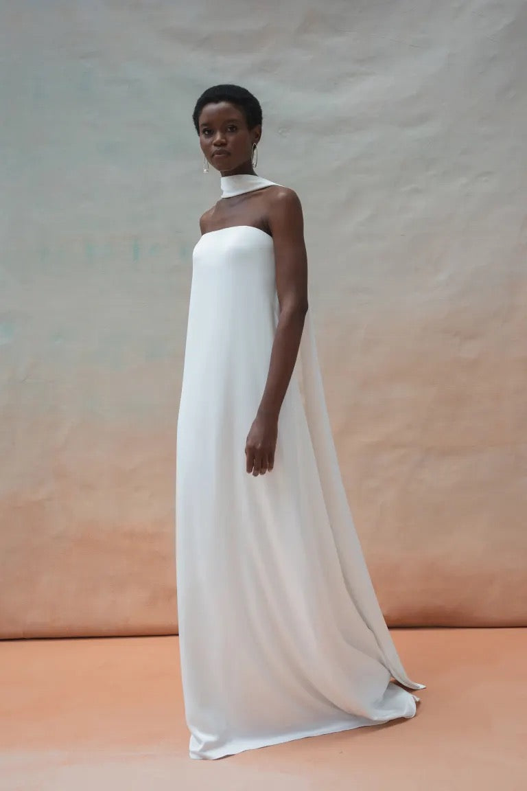 A person wearing the Celeste Wedding Dress by Jenny Yoo from Bergamot Bridal, which is a white, strapless, floor-length gown with a high neck scarf, poses against a neutral background, exuding elegant drama.