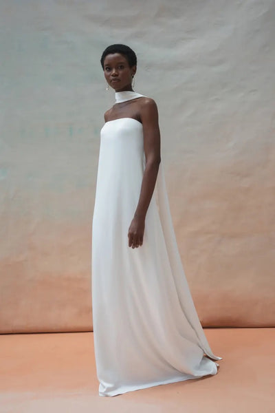 A person wearing the Celeste Wedding Dress by Jenny Yoo from Bergamot Bridal, which is a white, strapless, floor-length gown with a high neck scarf, poses against a neutral background, exuding elegant drama.