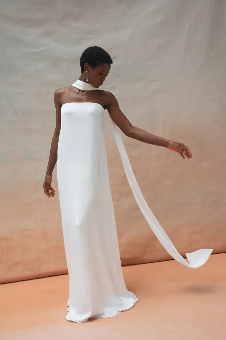 A person in the elegant Celeste Wedding Dress by Jenny Yoo from Bergamot Bridal, featuring a strapless white satin fabric and a long, dramatic scarf-like attachment, standing against a plain backdrop.