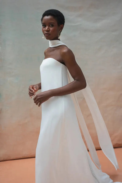 A woman in the strapless white Celeste Wedding Dress by Jenny Yoo from Bergamot Bridal, paired with a long scarf and set against a neutral background, exudes elegant drama.