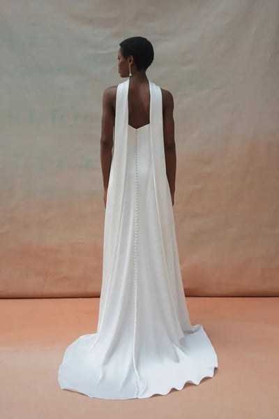 A person stands facing away, adorned in the stunning Celeste Wedding Dress by Jenny Yoo from Bergamot Bridal. The long, white gown features an elegant draped design and a stylish row of buttons down the back. Crafted from luxe satin fabric, it exudes a sense of graceful drama against the neutral, gradient backdrop.