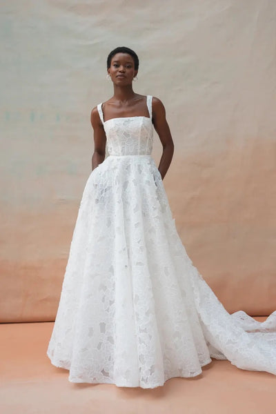 A person stands wearing the Charlie Wedding Dress by Jenny Yoo from Bergamot Bridal, a floor-length gown crafted from intricate white lace and floral organza fabric, featuring a patterned design and a train, photographed against a neutral backdrop.