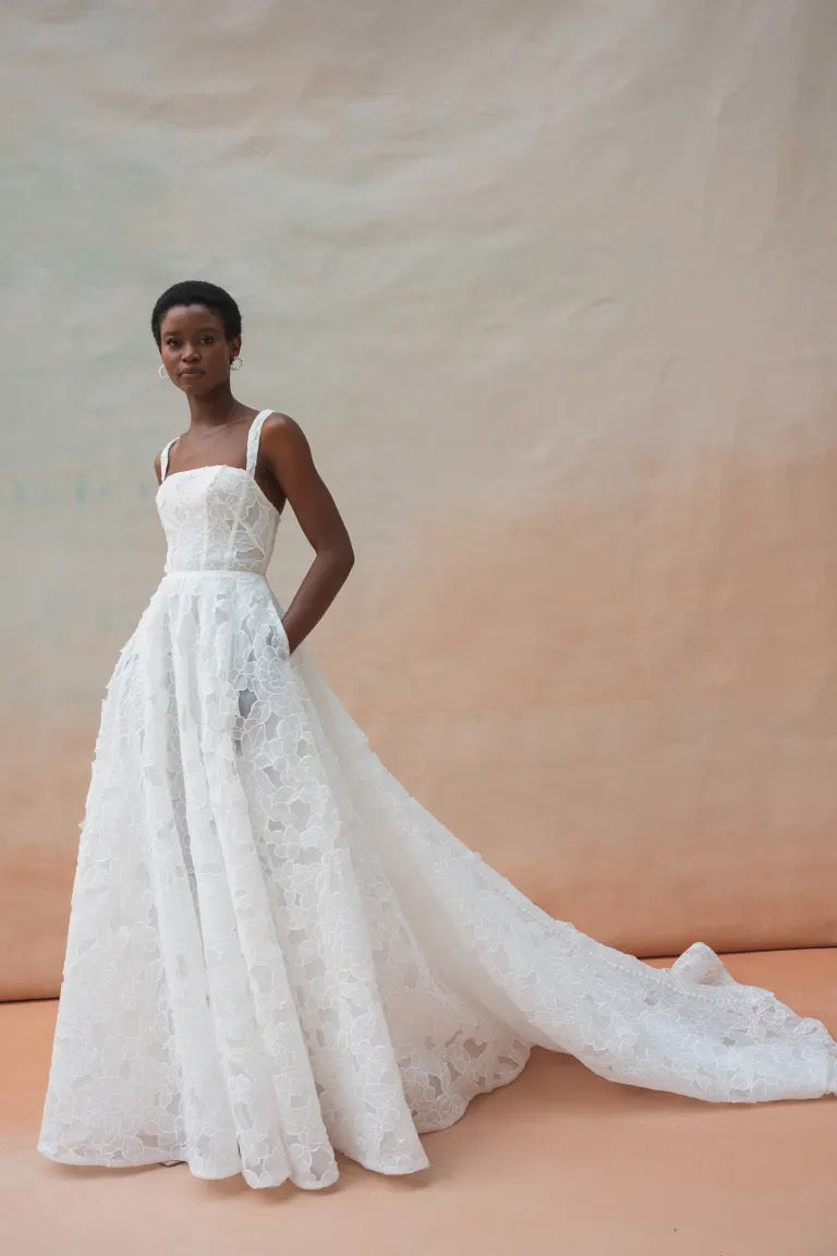 A model poses in a white lace Charlie Wedding Dress by Jenny Yoo, available at Bergamot Bridal. The gown features a sculpted corset and square neckline with delicate floral organza fabric and a full-length train, set against a muted background.