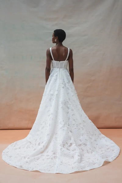 A person wearing the Charlie Wedding Dress by Jenny Yoo, available from Bergamot Bridal, is pictured from the back against a neutral background. The dress features white lace and floral organza fabric, complemented by a sculpted corset and a long train.